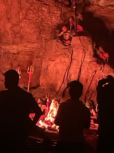 In Gollagutta, India, within a dimly lit cave adorned with red lighting and textured rock formations, a scene unfolds depicting a tableau of figures and symbolic objects. Silhouetted figures stand in the foreground, facing away from the viewer, their forms dark against the illuminated backdrop. In the middle ground, a figure, appearing to be a mannequin or statue, sits on a rock ledge wearing a red cloth headdress and loincloth, with arms outstretched. Nearby, a trident-like object, also red, is prominently displayed. Further into the cave, other figures and objects are partially visible, bathed in the red light, suggesting a theatrical or religious display. The environment is characterized by rough, uneven rock surfaces and a general atmosphere of dramatic presentation.