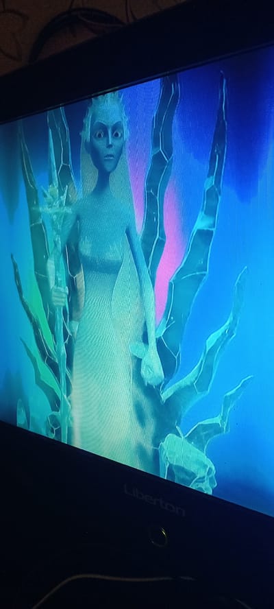 The image displays a still frame from a video game or animation, featuring a pale-skinned, humanoid character with wide, startled eyes and spiky, white hair, positioned centrally. The character appears to be a female figure, wearing a light-colored, flowing garment, and holding a staff-like object in her left hand. She is surrounded by abstract, crystalline or ice-like structures in shades of green and blue, which branch out around her. The background is a vibrant gradient of blue and pink. The screen itself is visible, with a black bezel and the "Liberton" logo subtly printed on the lower right corner. The image is captured from a slight angle, suggesting it is a photograph of a monitor or television screen. There is no indication of the city of Koviaghy, Ukraine, or any urban environment.
