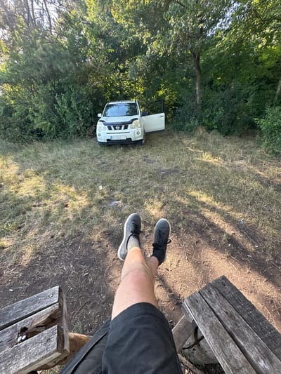 In the city of Velykozalissia, Ukraine, a white Nissan X-Trail SUV with license plate BX 8662 IC is parked on a dirt and grass clearing surrounded by dense green foliage and trees. The driver's side door of the vehicle is open. In the foreground, a person's legs, clad in black shorts and dark sneakers with gray socks, are visible, with their feet resting on two wooden benches. The lighting suggests it is daytime, with sunlight filtering through the trees and casting shadows on the ground.