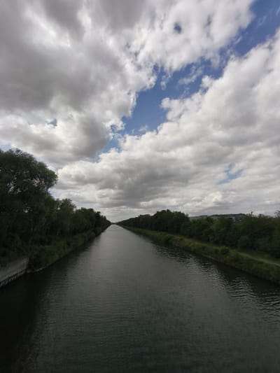 This is a vertical, eye-level, wide-angle photograph of a canal in Dourges, France. The canal runs from the foreground to the background, with its dark, rippling water filling the center of the frame. Lush green trees line both sides of the canal, with the trees on the left appearing thicker and closer to the camera, while the trees on the right are more distant and sparser. A narrow dirt path is visible along the right bank. The sky above is filled with dramatic, heavy clouds, with patches of bright blue sky visible in between. In the far distance, on the right bank, there is a large, modern industrial building with a grey facade. The overall impression is one of a calm, overcast day in a semi-industrial or suburban landscape.