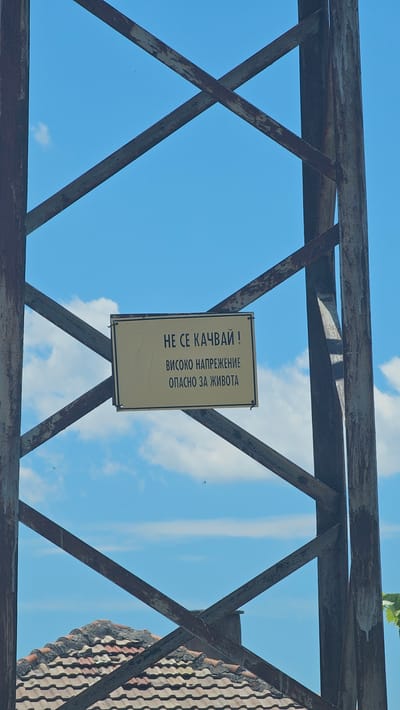 The image captures a rusty metal tower against a bright blue sky with scattered clouds. A rectangular yellow sign with black text is attached to the tower. The sign reads in Bulgarian: "НЕ СЕ КАЧВАЙ !" (Do not climb!) followed by "ВИСОКО НАПРЕЖЕНИЕ" (High voltage) and "ОПАСНО ЗА ЖИВОТА" (Dangerous for life). In the lower part of the image, a tiled roof of a building is visible. The scene appears to be in an outdoor, possibly rural or industrial setting, given the presence of the power tower.