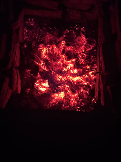 The image depicts the glowing embers of a fire within a stone fire pit at night. The embers are a vibrant red and orange, with some appearing brighter than others. The fire pit is surrounded by rough-hewn stones, some of which are illuminated by the fire's glow. The background is dark, suggesting it is nighttime and the surrounding area is unlit. The overall impression is one of warmth and intense heat emanating from the dying fire. The location is specified as Hancharski sel'ski saves, Belarus, though the image itself provides no specific clues to confirm this location.