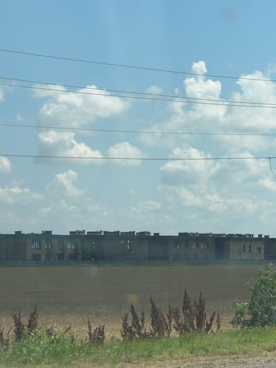 The image depicts a long, multi-story building with a flat roof, partially obscured by trees and a field in the foreground. The building appears to be commercial or industrial in nature, with numerous windows visible. The sky is a bright blue with scattered white clouds, and power lines are stretched across the upper portion of the frame. The foreground consists of a field of dry grass and some green vegetation. The overall scene suggests an outdoor setting, possibly on the outskirts of a city. The location is identified as Yelizavetinskaya, Russia, a rural settlement in Krasnodar Krai.