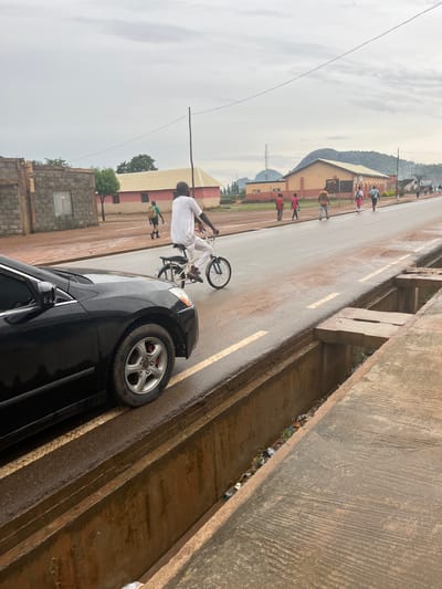 A black sedan is parked on the left side of a wet, paved road. The car is facing away from the viewer, with its front left wheel visible. A person wearing a white shirt and pants is riding a bicycle in the right lane of the road, also moving away from the viewer. Several other people are walking on the sidewalk to the right of the road. In the background, there are buildings and a hilly landscape under a cloudy sky. The scene appears to be a street in Sabojibi, Nigeria, with a drainage ditch running along the right side of the road, containing some debris.
