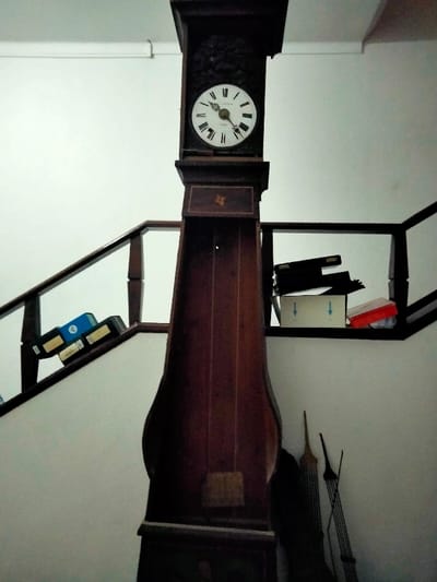 A tall grandfather clock stands against a white wall, positioned near a staircase with black railings. Books sit on the lower stair railing. On the upper stair railing, a box, a red object, and other items rest. Behind the clock, there are what appear to be long, slender components likely associated with the clock itself. The clock face is round with Roman numerals.