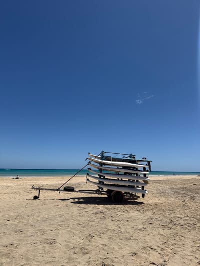 The image captures a sunny scene on a beach in Pájara, Spain. A tall, wheeled cart is loaded with numerous white surfboards or paddleboards, stacked horizontally. The cart casts a shadow on the light-colored sand. Beyond the cart, a calm, light blue sea stretches to the horizon, where it meets a lighter blue sky. The sky is mostly clear, save for a small, puffy white cloud. In the distance, people can be seen on the beach and in the water.