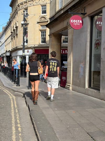 The image shows a street scene in Bath, United Kingdom, in front of a Costa Coffee shop. Two individuals are walking on the sidewalk. The man is wearing a black T-shirt with graphics, gray shorts, and socks. The woman is wearing a black outfit consisting of a top and shorts and brown knee-high boots. She has a small brown purse over her shoulder. Street furniture, including black bollards, is visible along the sidewalk. Several other individuals are also on the street and behind the bollards. The street has yellow lines painted on the ground. The buildings are primarily constructed with stone. A sign for Jones Westgate St. and Tree Good Foods can be seen.