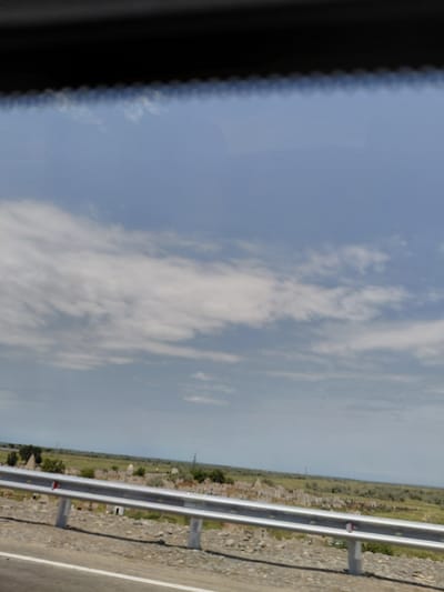 The image is a landscape view from a moving vehicle, likely a car, looking out through the windshield. The foreground is dominated by a metallic guardrail, typical of roadside safety barriers, with a white line marking the edge of a road beneath it. The ground beside the road is covered in gravel and sparse vegetation.

In the mid-ground, a vast, flat plain stretches out, dotted with numerous small, white structures that appear to be graves or mausoleums, suggesting the presence of a cemetery or historical burial site. Patches of green grass, shrubs, and some scattered trees are visible across the landscape. In the far distance, the horizon is marked by a hazy line where the land meets the sky.

The sky occupies the upper half of the image and is filled with a mix of white clouds and pale blue sky. The lighting suggests it is daytime, possibly late morning or early afternoon. The overall impression is of a rural or semi-rural environment.

While the specific location within Ақтүбек, Kazakhstan is not explicitly indicated in the image, the presence of such a large burial ground could be a significant landmark. The scene depicts a still, open landscape with a clear sky and evidence of human settlement or remembrance. There are no people or animals visible in the image.