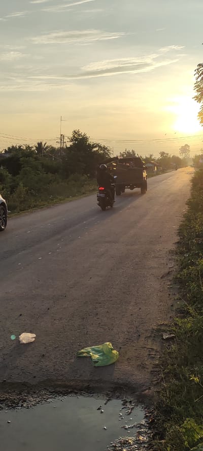 An outdoor, vertical, medium shot captures a rural road in Blangmajron, Indonesia during sunset. The sun is setting to the right of the frame, casting a golden glow across the sky and illuminating the scene with warm light. In the foreground, the asphalt road is marred by a puddle of murky water and scattered litter, including two plastic bags, one white and one green. A motorcycle, with a rider wearing a helmet and dark clothing, travels away from the viewer on the road. Behind the motorcycle, a truck, also carrying what appears to be cargo, moves in the same direction. The roadside is bordered by dense green vegetation and trees, with hints of scattered houses or buildings in the distance. Overhead power lines are visible against the pale, cloud-streaked sky. The overall impression is of a quiet, everyday scene on a rural road in Indonesia at dusk.