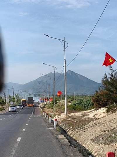 The image displays a multi-lane paved road extending into the distance in Tuy Phong 4, Vietnam, under clear daylight. A prominent mountain range with distinct peaks forms the background beneath a light blue sky. On the road, vehicles are visible moving away from the viewer, including a white sedan, a dark sedan, and a yellow semi-truck with a dark trailer. A truck further down the road emits dark exhaust smoke. The right side of the road is bordered by white and red concrete markers alongside a paved drainage channel. Beyond this, a series of streetlights with curved arms line the road, interspersed with poles flying red Vietnamese national flags featuring a yellow star, and at least one red flag displaying a yellow hammer and sickle. Sparse vegetation and sandy ground are present adjacent to the drainage, followed by a line of denser trees and shrubs. On the left side, utility poles with power lines run parallel to the road. The upper-left foreground contains a dark, blurry obstruction. No individuals are discernible.