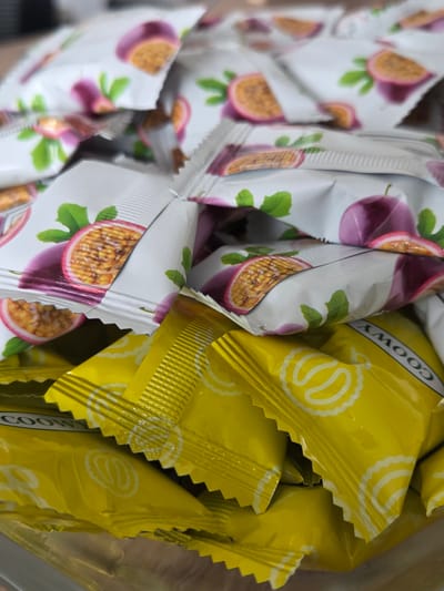 The image shows a pile of individually wrapped sweets. The top layer of sweets are wrapped in white packaging with illustrations of passion fruit and green leaves. The bottom layer of sweets are wrapped in bright yellow packaging with a white circular design and some text that appears to be "COOWY". The sweets are likely in a container or on a surface, but the background is blurred and out of focus, making it difficult to determine the exact setting. The image does not contain any information that would place it in Міханавіцкі сельскі Савет, Belarus.