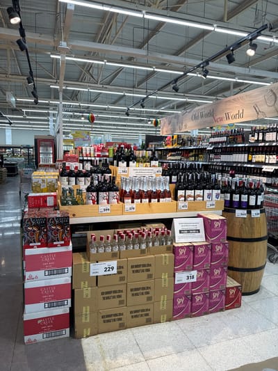 The image depicts the alcohol section of a grocery store, likely in Thong Sala, Thailand. The aisles are lined with shelves stocked with wine, spirits, and pre-mixed alcoholic beverages. Overhead, a banner distinguishes "Old World" and "New World" wines. There are many bottles of Mont Clair and Berri Estates. Displayed prominently at the end of the aisles are stacks of "Spy Classic" mixed drinks and "Castle Creek" dry red wine. Prices are displayed on nearby shelf-talkers. Boxes of Smirnoff vodka are stacked next to the Spy drinks. A wooden barrel rests atop the stacked wine. The scene includes metal shelves, overhead lighting, and a gray concrete floor.