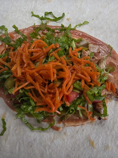 This is a top-down, close-up shot of a partially assembled wrap on a light-colored, textured surface, likely a countertop in the city of Ilyichbetovo, Russia. The wrap is made of a thin, white flatbread and is filled with a generous portion of ingredients. The filling consists of shredded lettuce, julienned carrots, chopped pickles, pieces of cooked meat (likely chicken or pork), and visible slices of tomato. A reddish-pink sauce, possibly a mayonnaise-based dressing or a sweet chili sauce, is spread over the flatbread and mixed with the filling. The ingredients are piled high in the center of the wrap, with some spilling over the sides. The lighting is bright and even, casting minimal shadows. The background is largely out of focus, emphasizing the food. No people or animals are present in the image.