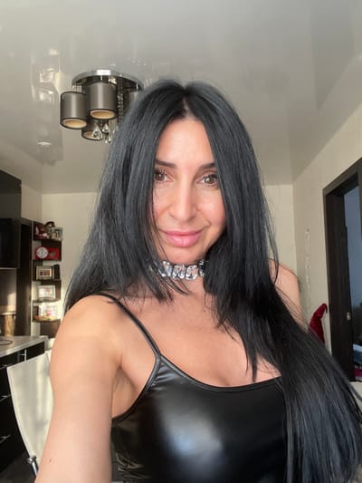 The image features a woman posing indoors, looking directly at the camera with a friendly, closed-mouth smile. She has long, straight, dark (likely black or very dark brown) hair, parted on the side, which falls over her shoulders and partially across her face. Her complexion is fair, and she has brown eyes and well-defined eyebrows. Her lips are a light pink shade.
She is wearing a form-fitting, shiny black top, possibly made of faux leather or a similar material, with thin spaghetti straps visible on her right shoulder. Around her neck, she wears a prominent, chunky silver-toned necklace or choker adorned with large, clear, rectangular or cushion-cut gemstones or crystals.
The background suggests an indoor setting, likely a home. To the left, a modern light fixture with three cylindrical, dark-colored shades is mounted on a light-colored wall. Below this, a dark-colored shelf or surface holds several small framed photos or decorative items and a dark mug. In the upper right corner, a portion of a dark-framed object, possibly a window or a piece of furniture, is visible. The lighting in the scene appears bright and even, indicating a well-lit room. No discernible text is visible in the image.