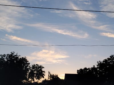 The image depicts a silhouetted rural landscape against a twilight sky. Two power lines stretch horizontally across the frame, dividing the sky into distinct sections. The upper portion of the sky is a pale blue, streaked with wispy white clouds that catch the fading sunlight. The lower portion of the sky transitions into warmer tones of yellow and orange near the horizon, indicating sunset or sunrise.

In the foreground, the dark silhouettes of trees and rooftops are visible, suggesting a village or scattered dwellings. On the left, dense foliage of trees dominates the lower edge. To the right of center, a house with a dark roof is prominent, with other smaller structures and trees also silhouetted against the sky. The overall impression is one of a quiet, end-of-day scene in a rural setting, likely in Cîrpești, Moldova, given the context. There are no discernible people or active interactions visible in the image. The environment appears calm and still.