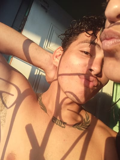 The image captures a close-up of a young person, seemingly preparing for a selfie with another person whose lips are visible in the foreground. The subject in focus has dark, curly hair and fair skin, with sunlight casting dramatic shadows across their face and upper body. They are shirtless, revealing a tattoo on their collarbone depicting a laurel wreath, and another tattoo on their neck that appears to be the number "2005". The background suggests an outdoor setting with a door and a wall, possibly in a residential area. The lighting indicates it is either sunrise or sunset. The overall impression is one of intimacy and personal connection, with the focus on the subjects' faces and their shared moment.