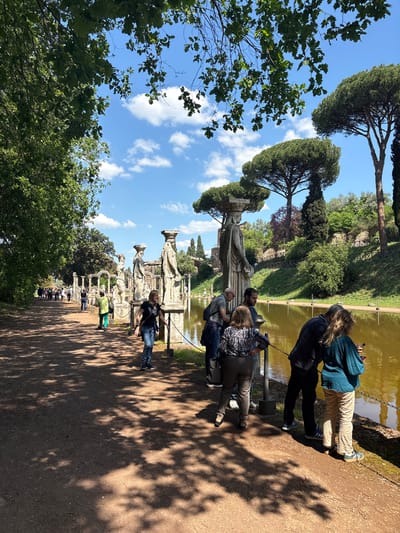 The image shows a park landscape with trees and a line of statues along a body of water, likely a lake or large pond, in the city of Tivoli, Italy. A dirt path runs parallel to the statues, and several people are present: some are standing by the water's edge, appearing to observe the scenery, while others are walking along the path in the distance. The sky is blue with some scattered clouds, and the lighting suggests it is daytime. The foreground includes trees, partially obscuring the view, and creating shadows on the ground. The statues themselves are light in color, potentially stone or concrete, and stand in a row alongside the water's edge. The scene has a relaxed, outdoor atmosphere typical of a public park or garden within a historical or tourist site.