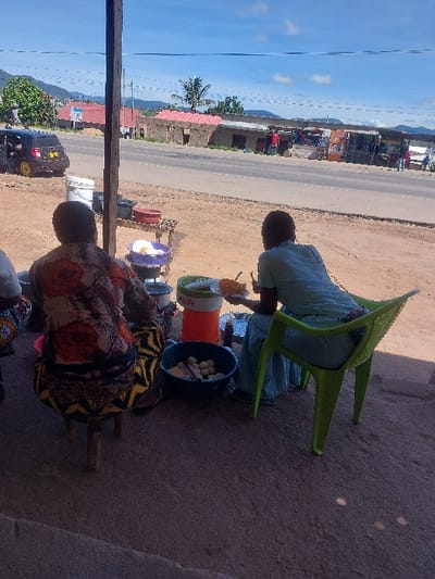 This image captures an outdoor scene in Kimamba, Tanzania, portraying an informal roadside food vending setup. In the immediate foreground, two individuals are seated with their backs largely towards the camera. On the left, a person sits on a low wooden stool, wearing a patterned top with red and orange floral designs over a dark background, and a yellow and black patterned lower garment. To their right, a second person is seated on a bright green plastic chair, dressed in light blue clothing, and is actively eating from a yellow bowl using a spoon.
Between these individuals, a small wooden stand holds various items including a white bucket and a purple basin containing several light-colored, ovoid objects. On the ground, an orange and white thermal flask is visible next to a dark blue basin filled with numerous rounded, light-colored food items. A wooden support pillar stands on the left, indicating a shaded area.
Across an asphalt road, a row of single-story buildings, some with red corrugated iron roofs and others with flat roofs, line the street. Multiple indistinct figures are present near these structures, suggesting commercial or residential activity. A dark-colored hatchback vehicle with a yellow rear wheel/hubcap is partially visible on the left side of the road. In the background, rolling hills or mountains are discernible beneath a clear blue sky with scattered clouds. Power lines stretch across the upper portion of the scene. The ground in the foreground and across the road is unpaved and dusty.