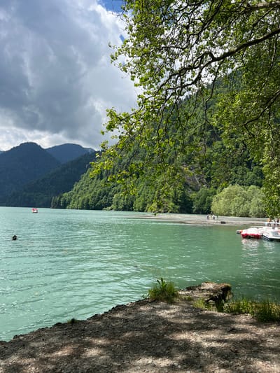 The image depicts a serene lake scene in Abkhazia. The lake's water is a vibrant turquoise, reflecting the cloudy sky above. Lush green mountains, densely covered with trees, form a dramatic backdrop to the lake. In the foreground, the shoreline is a mix of gravel and dirt, with some vegetation growing along the edge.
On the water, a small red and white boat is visible, and further out, another boat appears to be a paddleboat. Closer to the shore, on a sandy strip, a few individuals are standing. To the right, several white boats with red accents are docked. A portion of a tree with green leaves and branches is visible in the upper right corner of the frame, casting shadows on the foreground. The overall atmosphere is tranquil and natural.