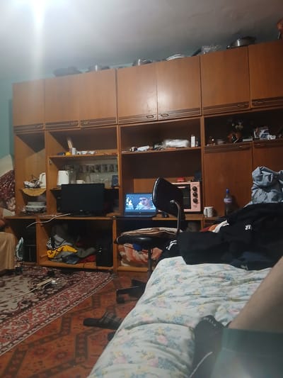 The image depicts a cluttered bedroom in an apartment in Devnya, Bulgaria. A large wooden wardrobe dominates the background, filled with various items including cookware on top, shelves with mugs and small appliances, and cabinets. In the foreground, a bed with a floral duvet is partially visible, with a person's legs and feet on it. A black office chair with a laptop is positioned in front of a desk area. On the desk, a laptop displays a man's face, a small pink microwave, and other miscellaneous objects. A red and orange patterned rug covers the floor, with a pair of plaid slippers near the chair. Clothes are strewn across the floor and on shelves. A bottle of "Devin" mineral water is visible on a shelf. The room appears to be dimly lit, with overhead lighting causing a bright glare in the upper left corner.
