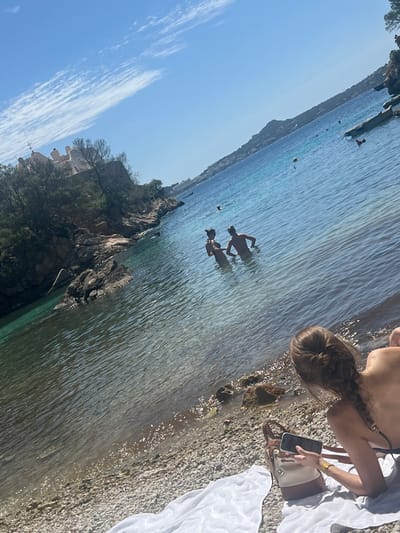 The image depicts a coastal scene in Peguera, Spain. Two individuals are seen waist-deep in the turquoise water, their figures slightly obscured by the water's surface. A woman stands on the rocky beach, facing the water, holding a smartphone. Next to her is a white towel, and a tan and brown satchel purse. The rocky coastline is visible along the left side of the image, with some trees and buildings on it. The sky is a clear blue with a few scattered clouds.