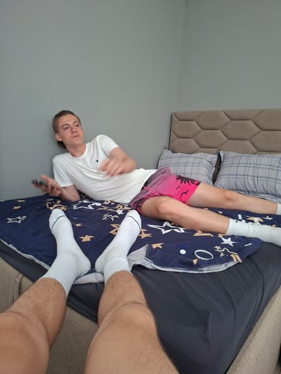 A young man with light skin and short brown hair is lying on a bed in a bedroom. He is wearing a white t-shirt and pink shorts with a palm tree print. His legs are extended, and he is wearing white socks. The bed is covered with a blue comforter with a star pattern and a gray blanket. There are two pillows on the bed, one with a gray and white plaid pattern and another with a blue and white checkered pattern. The background consists of a light gray wall and a portion of a beige headboard with a hexagonal pattern. The man is holding a black remote control in his right hand. His expression is neutral, and he appears relaxed. The lighting suggests it is daytime, and there are no indications of the weather. The scene is indoors, likely in a residential setting.