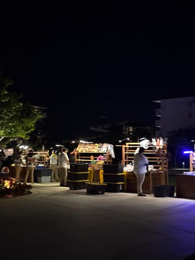 The image shows a night scene in Punta Sam, Mexico, likely at a resort or open-air market. Several people are visible, some appearing to be vendors at stalls displaying various goods, possibly souvenirs or crafts. The stalls are constructed with wooden shelving and are illuminated by ambient lighting. In the background, buildings with multiple windows are discernible, suggesting a resort setting. The ground is paved, and there are trees and some illuminated foliage in the foreground, adding to the tropical night atmosphere. The overall impression is of a lively evening market or gathering.