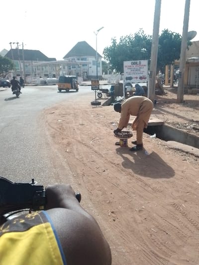 A man stands on a sandy roadside, tending to a tray of small brown goods, likely food. He's dressed in light brown clothing. Behind him, a drainage ditch runs along the roadside. A sign advertising "OBILO Technical Engineering" is visible in the background, near a building. Across the road, there's a gas station. A motorcycle and a yellow auto rickshaw move along the paved road. The photo is taken from the perspective of someone on another motorcycle, with the driver's arm and yellow-and-blue shirt visible.