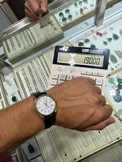 In Liwan District, China, a close-up shot focuses on a person's left forearm and hand, which is positioned over a calculator and jewelry displays. The calculator shows a "GRAND TOTAL" of "19000". The person is wearing a gold and white-faced wristwatch with a black leather strap. The background features multiple glass display cases filled with various pieces of jewelry, including necklaces, pendants, and bracelets, many of which appear to be made of gold, silver, and jade. The lighting suggests an indoor setting, likely a jewelry store.