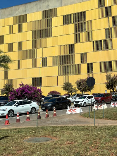 The image captures a street scene in Brasília, Brazil, with a tall yellow building facade on the left side. The facade is constructed with evenly spaced rectangular panels with horizontal slats, creating a grid-like pattern. A narrow sidewalk runs alongside the building, bordered by a grassy area. Cars are parked along the sidewalk, parallel to the building. Several traffic cones are placed between the cars and the grassy area. Street signs and small trees are visible in the background. The sky is blue in the top left.