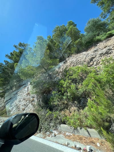 The image shows a view through a car windshield of a rocky hillside with dense green vegetation, including trees and bushes. The sky is clear and blue. A black car side mirror is visible in the lower left corner, reflecting a person's arm and hand. The scene appears to be a road passing by the side of the hill, possibly in a rural or coastal area. The lighting suggests it is daytime, with bright sunlight. The specific location in Mattinata, Italy is not discernible from the image itself.