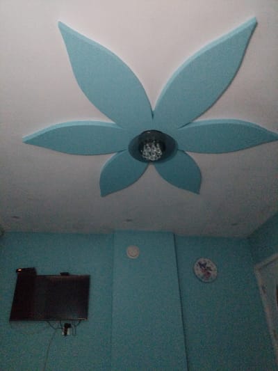 The image shows the interior of a room. A light blue, stylized flower-shaped decoration is mounted on the ceiling, with a small, multi-faceted light fixture at its center. Below the ceiling, the walls are painted a textured turquoise color. Mounted on the left wall is a flat-screen television. To the right of the television, on the same wall, is a circular clock with a floral design. Another circular object, possibly a vent or a light fixture, is visible higher on the wall. The image is taken from a low angle, looking upwards towards the ceiling. There is no visible indication of the city of Rakitovo, Bulgaria within the image itself.