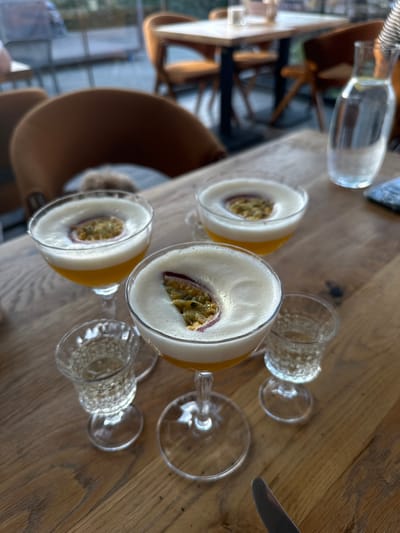 In Ivano-Frankivsk, Ukraine, on a wooden table, are three cocktails. Two of the cocktails are in coupe glasses, filled with an orange liquid topped with a frothy white foam, and each garnished with a slice of passion fruit. A third coupe glass, partially visible, also appears to contain a similar cocktail. Two small shot glasses, one on each side of the central coupe glass, are filled with a clear liquid. In the background, blurred images of chairs and tables suggest a restaurant or cafe setting. A glass carafe of water with lemon slices is visible on the right side of the image. The lighting suggests it is daytime, possibly late afternoon.