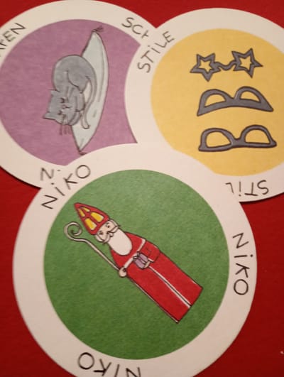 The image displays three circular cards arranged on a red surface. The card at the top left features a purple background with a drawing of a gray cat lying on its side. Text in black letters is visible on the outer white border: "PEN" on the left and "SCH" on the top.

The card at the top right has a yellow background and shows two pairs of glasses and two stars drawn in dark gray. Text on the outer white border reads "STILE" in black letters.

The card at the bottom has a green background and depicts a drawing of Saint Nicholas wearing a red robe, a red and yellow bishop's mitre, and holding a staff. Text on the outer white border reads "Niko" on the left, and "NIKO" on the right, with "STIL" upside down at the top right.

The cards appear to be game pieces or decorative items. No specific location in Rosenheim, Germany is depicted or mentioned in the image.