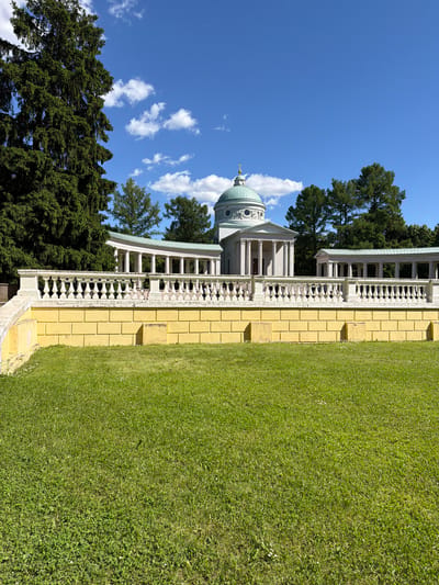 The image depicts an exterior view of a classical-style building with a domed roof, situated within a manicured park or garden. The building features white columns, ornate balustrades, and a pale yellow facade. A lush green lawn occupies the right side of the frame, contrasting with the architectural elements and the vibrant blue sky dotted with scattered white clouds. Tall, dark green evergreen trees frame the left side of the scene. The perspective is from a low angle, looking up towards the building and sky. The sunlight suggests it is daytime, likely morning or afternoon, with clear weather. There is no visible text in the image, and no people or animals are present. The scene conveys a sense of serenity and historical grandeur.
