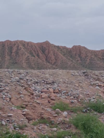The image shows a landscape view in the city of Kaji-Say, Kyrgyzstan. In the foreground, there is a rocky, uneven terrain with scattered green vegetation. The middle ground features a higher embankment of rocks and dirt, with more sparse vegetation. In the background, a range of eroded, reddish-brown mountains stretches across the horizon under a cloudy, overcast sky. There are no people or distinct structures visible in the image.