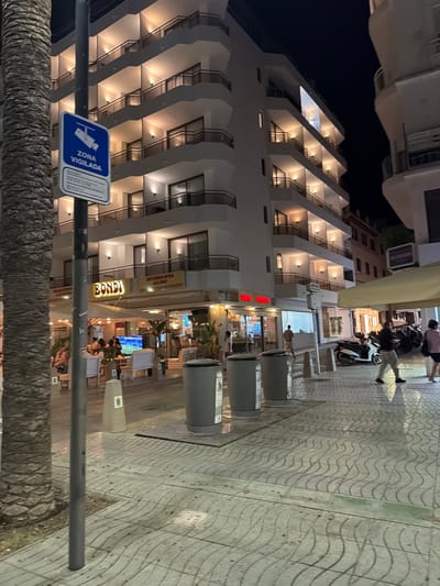 The image shows a street scene at night in Sant Antoni de Portmany, Spain. The main subject is a multi-story hotel with illuminated balconies, which dominates the left side of the frame. In front of the hotel, there's a sidewalk with a wavy pattern. Several people are visible on the sidewalk, some walking, some standing, and some seated at an outdoor restaurant or bar.

Objects present include several large trash bins on the sidewalk, a motorcycle parked near the hotel entrance, and a sign with a blue background displaying a camera icon and the text "ZONA VIGILADA," which translates to "Guarded Area." Another sign on the hotel reads "BONDIB." A yellow sign attached to the hotel states "KITCHEN OPEN ALL DAY."

The lighting suggests it is nighttime, with artificial lights illuminating the hotel and parts of the street. The overall atmosphere appears to be one of urban nightlife or evening activity. The context provided indicates this is in Sant Antoni de Portmany, Spain.