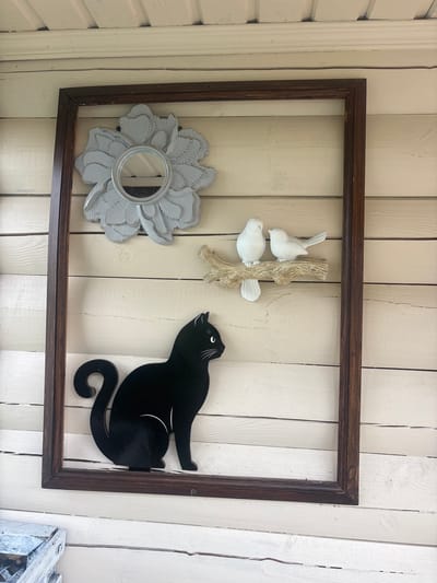 A dark brown wooden frame hangs on a weathered, pale yellow wooden wall, possibly the exterior of a building in Лотос Снт (марьино), Russia. Inside the frame, on the wall, are several decorative items. At the top left, a light gray decorative mirror shaped like a flower is mounted. To the right of the flower mirror, a faux branch with two small white bird figures is attached to the wall. Below the birds, a black silhouette of a cat is affixed to the wall, positioned at the bottom of the frame. The wooden wall exhibits horizontal planks with visible grain and some signs of wear. In the lower left corner, a portion of a white, weathered object, possibly a box or planter, is visible.
