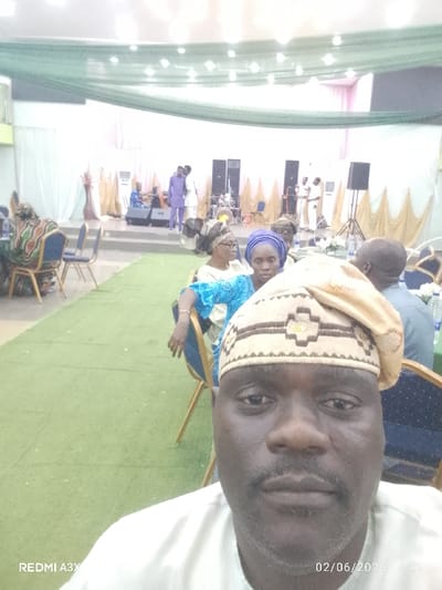 A selfie captures a man in a beige hat and light-colored shirt at an event. Behind him, several people are seated at tables on green flooring. A raised stage features musicians, speakers, and a backdrop of white fabric with gold accents. Speakers are positioned on either side of the stage. The ceiling is adorned with green and white fabric draping. The date "02/06/2026" is visible in the lower right corner.