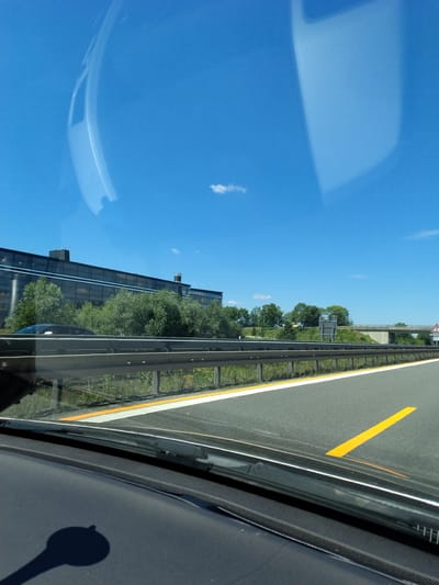 The image captures a view from inside a vehicle, likely on a highway, with a clear blue sky overhead and a few scattered clouds. In the distance, a modern-looking building with extensive glass windows is visible, surrounded by lush green trees. A highway guardrail runs parallel to the road, with a yellow line marking the lane. The scene appears to be set in the city of Hengersberg, Germany, given the context provided. There are no people or explicit actions depicted, but the presence of the guardrail and road markings suggests ongoing traffic. The overall impression is one of a bright, clear day, possibly during a journey through the German countryside or outskirts of a city.