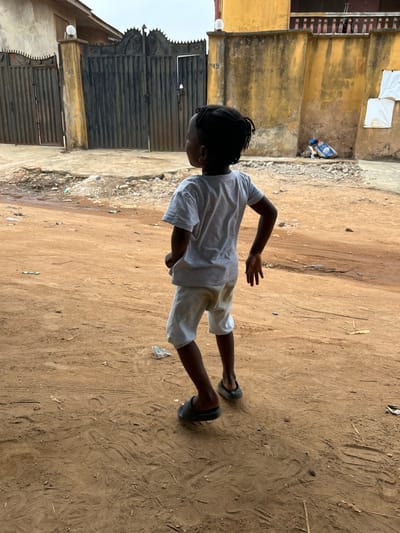 The image depicts a young child standing on an unpaved, reddish-brown dirt road in Ashipa, Nigeria, viewed from behind. The child has dark skin, dark hair styled back, and wears a light grey t-shirt, soiled white shorts, and dark slip-on sandals. A small gold earring is visible on the left ear. The child's right arm is slightly bent at the elbow, and the left hand is positioned near the hip. The child is looking towards the viewer's left. The road surface is uneven, featuring scattered small rocks, dirt, and minor debris.

In the background, to the left, a light-colored building is partially visible, featuring two dark grey metal gates. House numbers "48" and "49" are affixed to the adjacent walls. Dome-shaped light fixtures are mounted on pillars between and to the right of these gates. To the right, a two-story ochre-yellow building is visible, with a dark railing on its upper floor. A matching yellow wall extends from the building, against which a pile of debris, including a blue sack, is present on the ground. Two white rectangular objects are affixed to this yellow wall. The sky overhead is overcast.