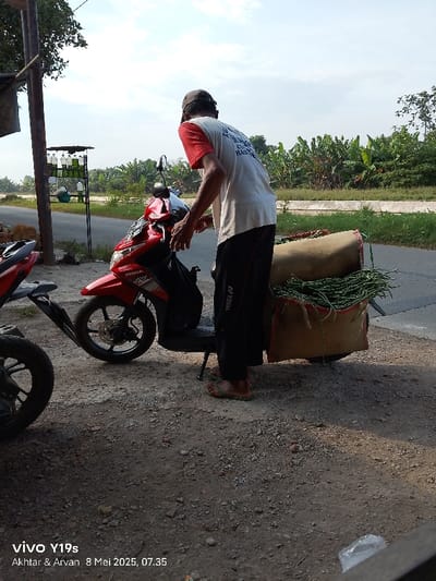 A man is standing next to a red Honda motorcycle. He is wearing a white shirt and black pants. The motorcycle is carrying a burlap bag full of green vegetables. The scene appears to be on a paved road. The man is located in Kecamatan Anjatan, Indonesia, at latitude: -6.42, longitude: 107.978. The environment is rural, with vegetation on either side of the road. There is another motorcycle partially visible in the background.