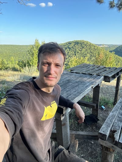 The media file shows a man taking a selfie outdoors. He is sitting on a rustic wooden picnic table in what appears to be a natural setting with lush green trees and rolling hills in the background. The sky is clear and blue, with a few white clouds visible, suggesting a pleasant day. The man is wearing a dark long-sleeved shirt with a yellow graphic design on it, and he has a light beard. His expression is relaxed and he is looking directly at the camera.
The picnic table is made of weathered wood, and there is some debris around it, including a discarded can on the ground. The overall scene suggests a casual outdoor break, possibly during a hike or a visit to a scenic spot. The time of day appears to be daytime, likely late morning or afternoon, given the bright sunlight. There is no visible text in the scene, other than what appears on the man's shirt, which is partially obscured but seems to have "THE ORIGINAL" written on it. The location context is Malozalissia, Ukraine.
