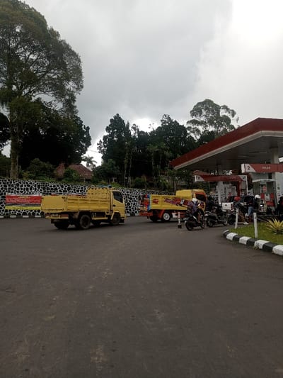 A gas station with a red roof is busy with motorbikes and people. Two yellow trucks are parked nearby. The scene is set on a cloudy day with lush green trees surrounding the area. A stone wall with signs on it separates the station from the greenery. The asphalt ground covers the foreground and leads into the bustling area.