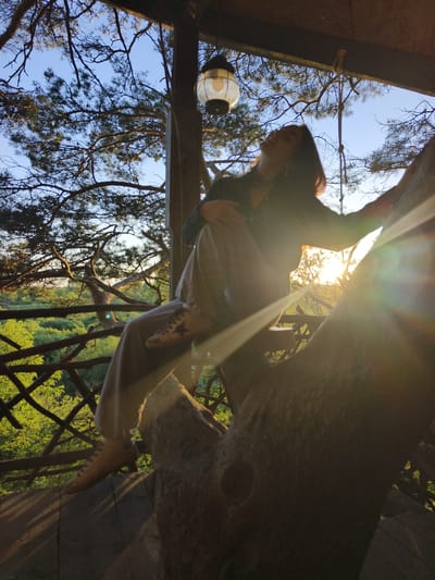 A silhouette of a woman is visible in a treehouse setting, with the sun setting behind her, creating a lens flare effect. She is sitting on a thick tree branch, with her head tilted back and eyes closed, appearing relaxed. She is wearing light-colored pants and a dark top. The treehouse structure is made of wood, with railings and a light fixture hanging from the ceiling. The surrounding environment is lush and green, with trees and foliage visible in the background, suggesting a natural setting outside the city. The overall mood is tranquil and serene. The location is the city of Onochino, Russia.