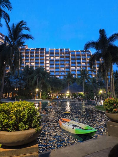 The image depicts a resort pool area at dusk in Tianya District, China. A large, multi-story hotel with illuminated windows and decorative string lights is visible in the background, surrounded by palm trees and lush vegetation. In the foreground, the pool water reflects the lights from the hotel and sky, with several people swimming and relaxing. A brightly colored kayak, with orange, green, and white sections, floats on the surface of the pool. Two large potted plants, one with dense green foliage and the other with red flowers, flank the pool area. The scene suggests a resort setting designed for leisure and recreation.