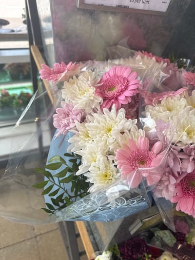 A close-up shot captures several wrapped bouquets featuring white and pink chrysanthemums. Some bouquets show only the flower heads, while others reveal the supporting greenery. The bouquets are encased in clear plastic wrap, with one bouquet wrapped in a light blue decorative wrapper. The image is taken at an angle, offering a partial view of the shelf or surface on which the flowers are arranged.