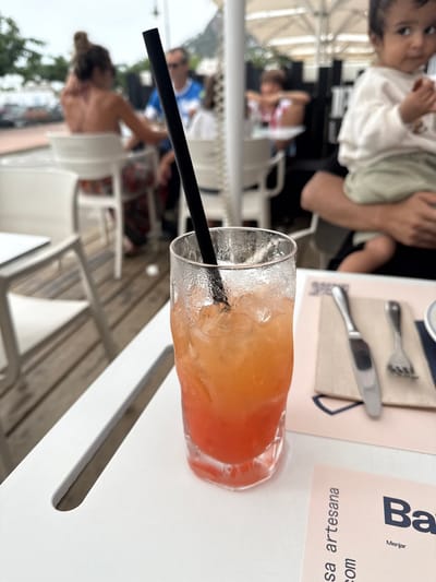 The image depicts an outdoor dining area in L'estartit, Spain, under an overcast sky. In the foreground, a heavily condensed clear glass holds a layered iced beverage, transitioning from reddish-pink at the bottom to orange-yellow at the top, containing numerous ice cubes and a black straw. This glass rests on a white outdoor table. To its right, a light pink paper placemat is set with a metal knife and fork, alongside a partially visible white circular dish. Below the placemat, a pink paper menu or advertisement partially displays the text "sa artesana" and "Bar Menjar."

In the mid-ground to the right, an adult's arm supports a young child with dark hair, wearing a light-colored long-sleeved top and green shorts, facing forward. Further into the mid-ground, several blurred individuals occupy white outdoor chairs around white tables on a wooden deck. Noteworthy figures include a woman with reddish-brown hair tied up, viewed from the back, revealing shoulder tattoos, and a man in a blue shirt wearing sunglasses. The distant background shows indistinct vehicles on a street, with hills or mountains under the cloudy sky.