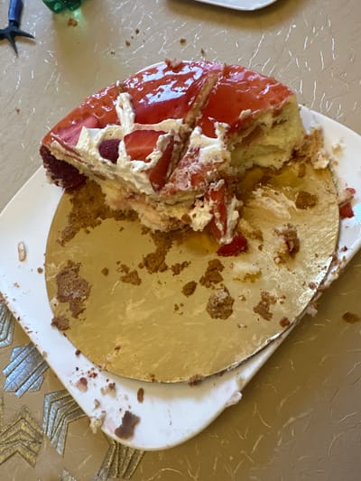 The image displays a partially consumed strawberry and cream cake, resting on a circular golden cake board placed centrally on a white, square plate with slightly rounded corners. Approximately half of the cake remains, showing layers of light-colored sponge, white whipped cream, sliced strawberries, and at least one raspberry, all covered with a reflective red fruit glaze. The exposed golden cake board and parts of the white plate are soiled with cake crumbs and smeared cream/red fruit residue, indicating recent consumption. The plate rests on a light tan-colored table surface or tablecloth that exhibits a subtle, reflective, geometric pattern. In the upper left background, a pair of black-handled pliers and a translucent green plastic bottle are partially visible. This scene is located in the city of Marcoussis, France.
