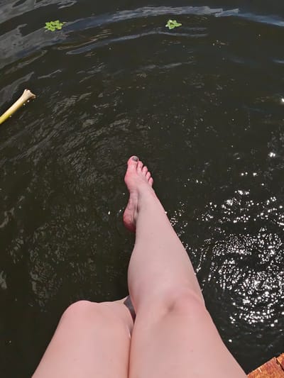 A person's bare legs and feet are visible, submerged in dark water. The water has ripples and reflections of sunlight. Two small green plants float on the surface of the water. A yellow object, possibly a stick or a piece of wood, is partially submerged on the left side of the image. The person appears to be sitting on a wooden surface, which is visible at the bottom right of the frame. The scene is set outdoors, likely in a natural body of water.